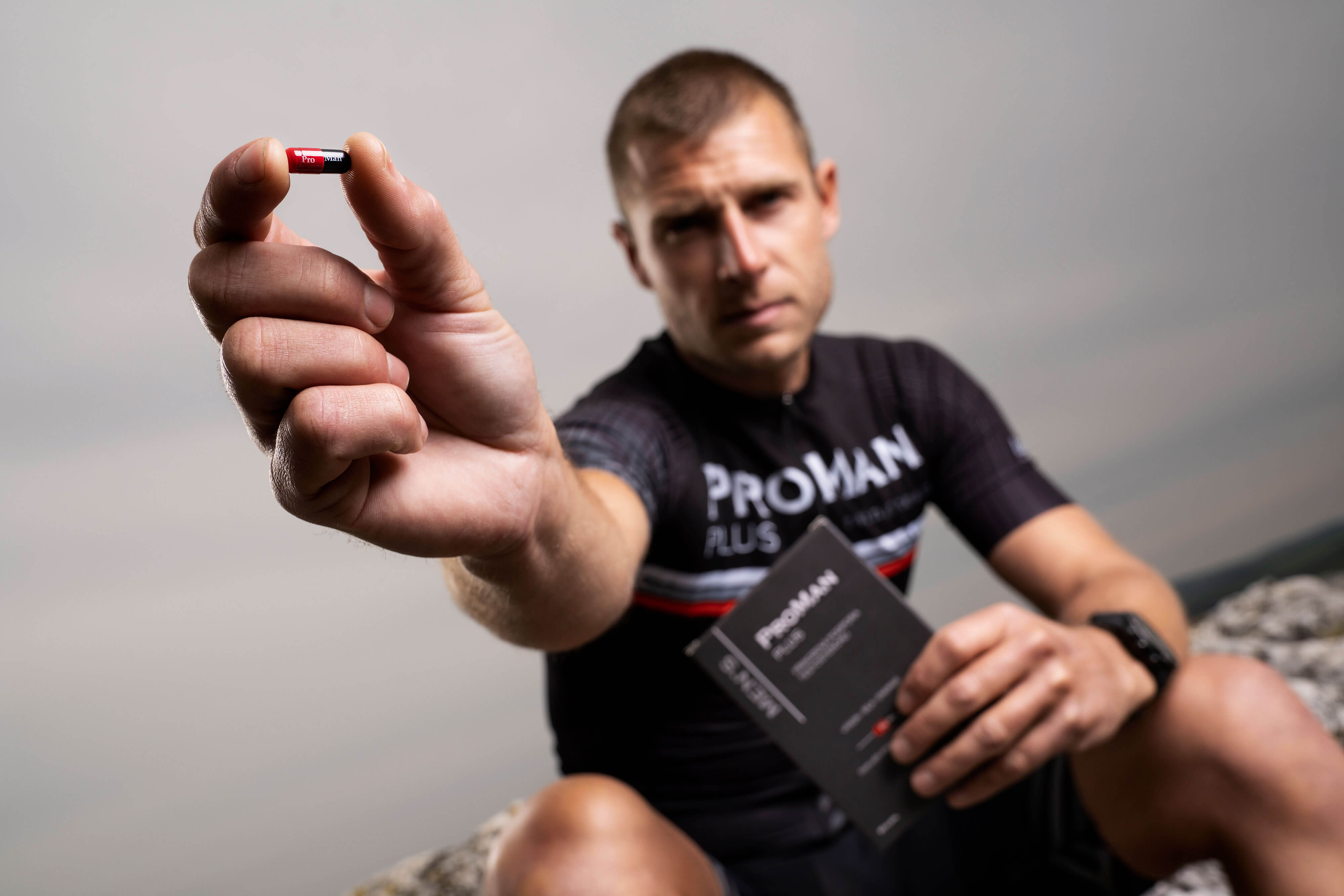 Natural Pharmaceuticals ProMan® Plus Mees spordirõivais „ProMan Plus Racing Team” hoiab väljasirutatud käes toidulisandi „ProMan® Plus” kapslit (punane-must). Teises käes hoiab ta toote pakendit. Taust on ähmane, väljas, hall taevas ja kaljud taustal.