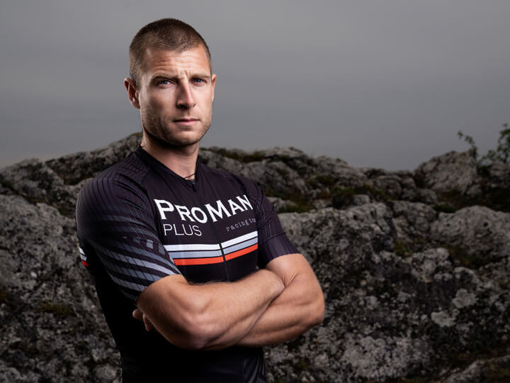 Natural Pharmaceuticals ProMan® Plus Mees spordirõivais, millel on kiri „ProMan Plus Racing Team”, seisab käed risti rinnal kaljuse maastiku taustal. Pilt on tehtud pilvise ilmaga, mis rõhutab modelli tõsist ilmet.